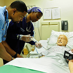 Nursing students in scrubs practice on a mannequin in a hospital room. 